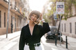 © PhotoBook - Adorable stylish woman with short hairstyles is touching her hair and posing to camera on city blurred background