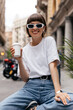 © PhotoBook - Stylish fashionable woman in sunglasses holding coffee and smiling to camera while resting outdoor