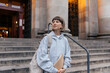 © PhotoBook - Romantic charming stylish teenager girl with short hair wearing grey shirt is holding backpack and notes stands in front of the university after classes
