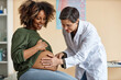 © Seventyfour - Medium full shot of senior Caucasian female gynecologist using hands for examining cheerful pregnant African American womans uncovered abdomen in clinic