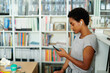 © Just images - young afro black woman casually engaged with her smartphone, earbuds in, in a relaxed home environment with a bookshelf backdrop suggesting a blend of leisure and technology.