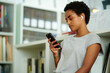 © Just images - A contemplative young afro black female student  engaging with her smartphone, earbuds in, against a calm student library, embodying the quiet focus of the digital age.