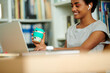 © Just images - A smiling black woman enjoys a coffee break while working using laptop at office, her satisfaction evident in her relaxed demeanor and the comfort of her home office.