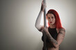 Waist up shot of red-haired young Caucasian woman with arm tattoos looking at camera and holding on to pole standing in studio backlight