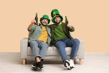 Wall Mural - Happy young men in leprechaun hats with green beards holding bottles of beer and sitting on sofa near beige wall. St. Patrick's Day celebration