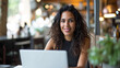 © Kowit - Cheerful young woman with natural hair smiles while working on her laptop in a modern cafe setting, radiating positivity and productivity.