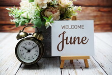 Welcome June Text Message Written On Paper Card With Wooden Easel And Alarm Clock With Flower In Metal Vase Decoration
