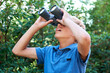 Boy, child with binoculars and surprise, search in nature for learning and fun, forest and adventure at summer camp. Young camper, leaves and trees, explore environment outdoor and wow for discovery
