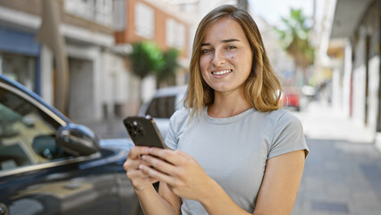 Canvas Print - Cheerful young blonde woman happily touching her smartphone, engaged in a digital conversation on a sunny city street