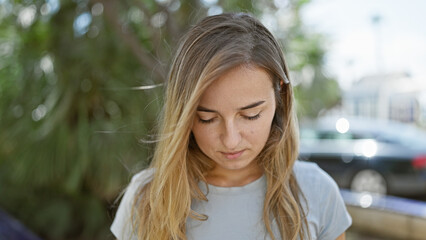 Poster - Unseen beauty, portrait of a young, blonde woman, striking a cool, casual pose, looking down in thought - seriously engrossed in the lush green park on this sunny day.