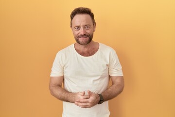 Wall Mural - Middle age man with beard standing over yellow background with hands together and crossed fingers smiling relaxed and cheerful. success and optimistic