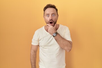 Wall Mural - Middle age man with beard standing over yellow background looking fascinated with disbelief, surprise and amazed expression with hands on chin