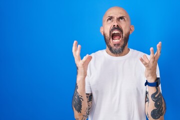 Wall Mural - Hispanic man with tattoos standing over blue background crazy and mad shouting and yelling with aggressive expression and arms raised. frustration concept.