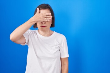 Wall Mural - Middle age hispanic woman standing over blue background covering eyes with hand, looking serious and sad. sightless, hiding and rejection concept