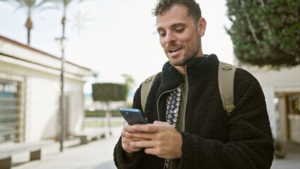 Poster - Smiling bearded young man using smartphone on a sunny city street, depicting urban life and technology.