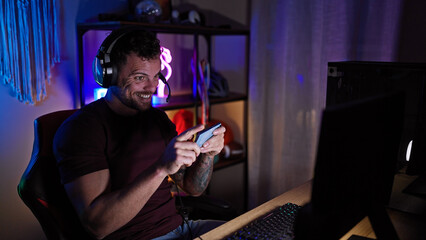 Wall Mural - Handsome hispanic man gaming at night in a dark room with colorful led lights wearing headphones and smiling.