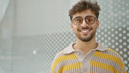 Poster - Confident young arab man, radiating joy and positivity, is standing outdoors on a city street, resplendently handsome in his sunglasses, his contagious smile lighting up the urban background.