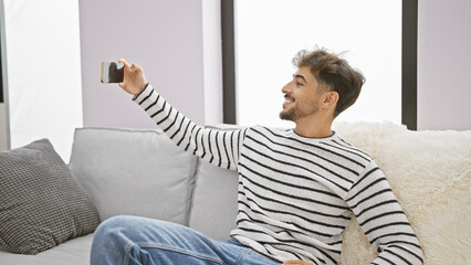 Sticker - Carefree, young arab man at home, happily enjoying the moment, expressing joy, taking a selfie on his smartphone while sitting on the sofa in living room