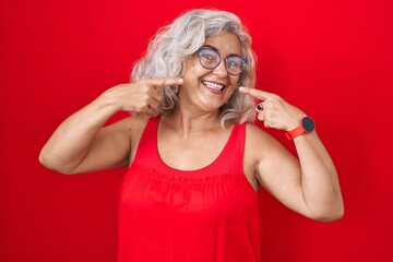 Sticker - Middle age woman with grey hair standing over red background smiling cheerful showing and pointing with fingers teeth and mouth. dental health concept.