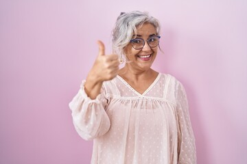 Poster - Middle age woman with grey hair standing over pink background doing happy thumbs up gesture with hand. approving expression looking at the camera showing success.