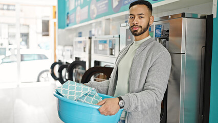 Wall Mural - Young hispanic man holding basket with clothes at laundry facility