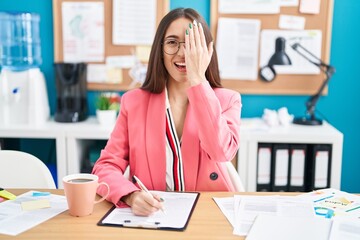 Wall Mural - Young hispanic woman working at the office wearing glasses covering one eye with hand, confident smile on face and surprise emotion.