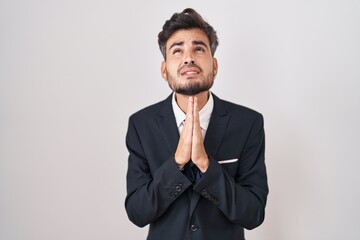 Sticker - Young hispanic man with tattoos wearing business suit and tie begging and praying with hands together with hope expression on face very emotional and worried. begging.