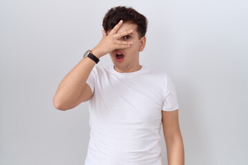 Poster - Young non binary man wearing casual white t shirt peeking in shock covering face and eyes with hand, looking through fingers with embarrassed expression.