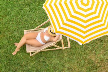 Woman in a white bikini sitting on deck chair under yellow umbrella  on the green grass sunbathes at summer day. Top view, drone, aerial view.