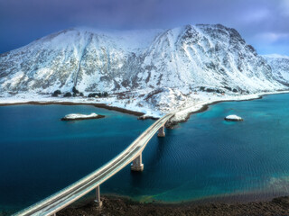 Wall Mural - Aerial view of bridge over the sea and snowy mountains in Lofoten Islands, Norway. Top drone view of bridge in cold winter day. Wintry landscape with blue water, rocks in snow, road, cloudy sky. 