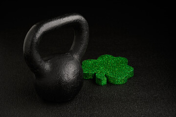 Wall Mural - Holiday fitness and Happy St. Patick’s Day, green glitter shamrock shape with a black iron kettlebell on a black background
