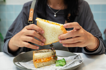 Sticker - Closeup of a woman holding and eating a sandwich with eggs and cheese and cucumber on a plate