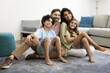 © fizkes - Happy middle aged couple of parents holding two sibling kids on lap, sitting on floor at cozy home, looking at camera away, smiling, laughing, enjoying family leisure, warm relationship