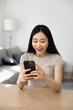 © fizkes - Happy Asian gen Z winner girl looking at smartphone screen, laughing, gasping, shouting for joy, feeling cheerful, excited about success, achievement, luck, saying yes, sitting at home workplace table