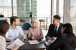 Positive senior business leader woman meeting with younger colleagues at negotiation table, offering ideas for brainstorming. Senior boss talking to listening coworkers, instructing team