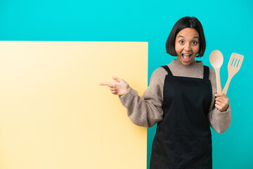 Wall Mural - Young mixed race cook woman with a big placard isolated on blue background surprised and pointing finger to the side