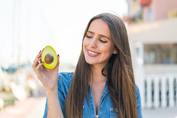 Wall Mural - Young woman holding an avocado at outdoors with happy expression