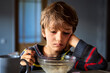 Dissatisfied Caucasian young boy is having breakfast while sitting in front of transparent bowl of porridge in dimly lit kitchen.