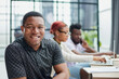 © Katsiaryna - young African American man in casual clothes posing at the camera while sitting at the table against the background of his colleagues