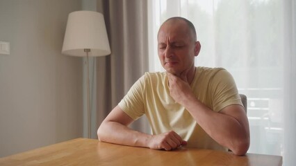 Wall Mural - Unhappy man coughing while sitting at table in living room at home. Sick with a cold or flu male feels bad, holds his hand to his chest. He has a bronchitis. 