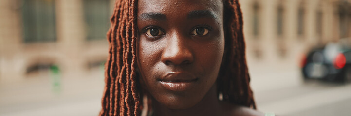 Wall Mural - Close-up of beautiful woman with African braids raising her head and looking at the camera with smile on the building background, Panorama.