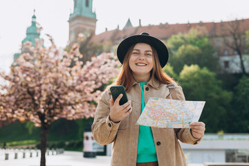 Wall Mural - Attractive young female tourist is exploring new city. Redhead 30s girl holding a paper map and smart phone in Krakow. Traveling Europe in spring. High quality photo