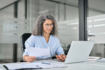 Busy mature business woman working in office looking at laptop holding document. Older professional hr manager, lawyer or notary checking financial tax report account in office. Business consultancy.