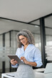 © insta_photos - Mature happy businesswoman bank manager, older female corporate executive holding digital tablet standing at work. Busy middle aged professional business woman using tab computer in office. Vertical