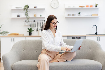 Focused caucasian woman dressed in casual attire typing on wireless laptop while sitting on comfy sofa. Attractive brunette female freelancer doing online work while staying at home.