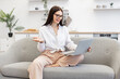 © sofiko14 - Charming female freelancer sitting on couch and using modern laptop for video conference at home. Confident brunette woman in talking on webcam and gesturing with hands.