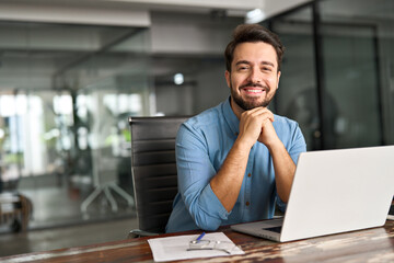 Happy professional business man company employee, young entrepreneur, smiling latin businessman working on laptop computer technology looking at camera working in office sitting at desk, portrait.