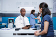 © DC Studio - In hospital room, African American male doctor in a lab coat explains healthcare and medicine to a Caucasian woman. A female nurse performs a medical checkup on a small girl in the background.