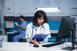 Female doctor preparing for medical consultations using a clipboard and computer. Image shows caucasian woman wearing a lab coat and writing down notes in a clinic office.