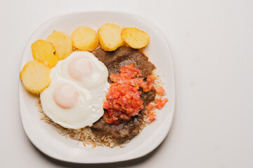 Top view of authentic, traditional Bolivian meal: Silpancho, made of beef, potatoes, rice, fried eggs, tomato and onion salad. close-up on white plate on white background.copy space